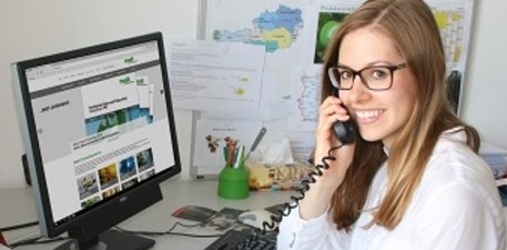 Eine blonde Frau mit Brille sitzt an einem Schreibtisch, schaut auf den Computer und telefoniert.